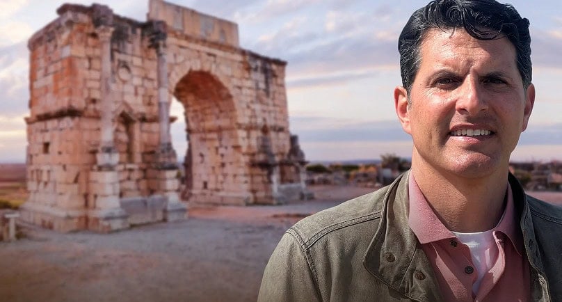 A man stands in front of an ancient stone archway in an open area, with blue sky and clouds in the background.