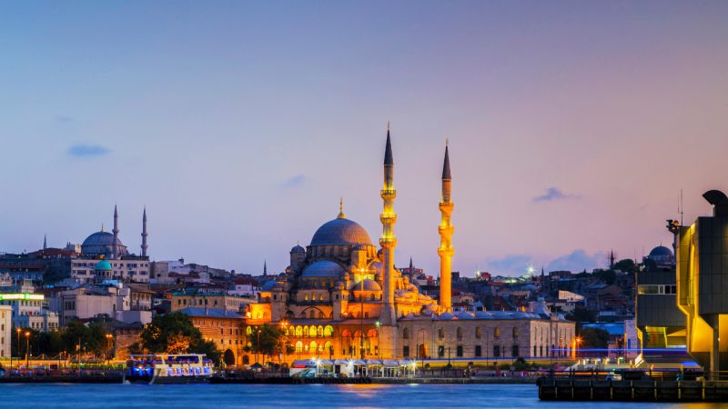 The illuminated New Mosque and its minarets on the Istanbul waterfront at dusk.