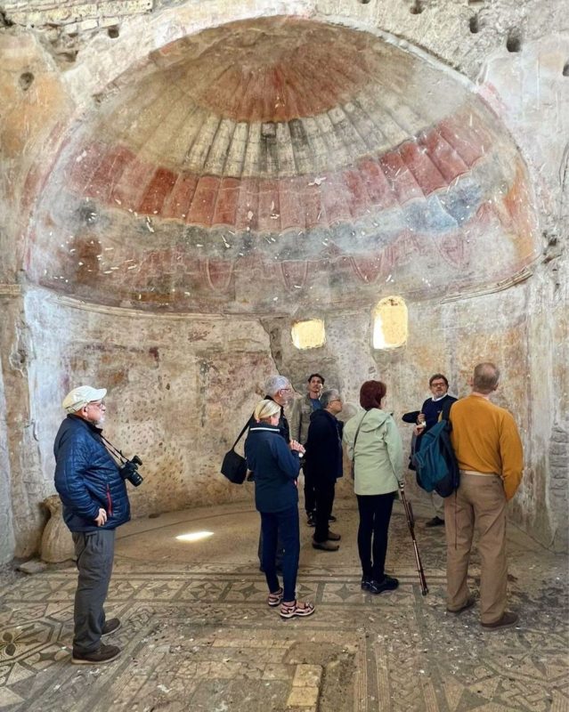 A group of tourists listen to a guide inside an ancient stone chamber featuring a domed ceiling with faded, arched frescoes.